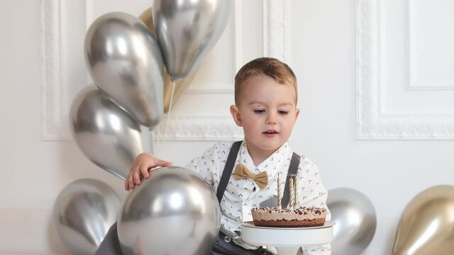 Little boy celebrating birthday, child has a B-day party. Boy blowing candles on the birthday cake. Celebration, white minimalist interior.
