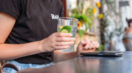 Woman at cafe drinking water and using mobile phone. Hand holding lemonade on cafe background. Glass of Cold drinks. Street front view, girl waiting for her order