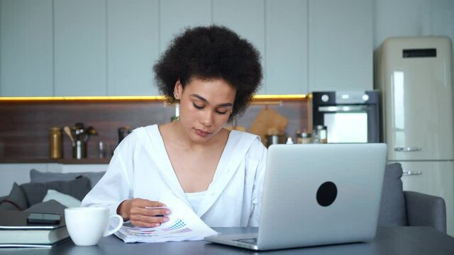 Young employee works at laptop remotely, analyzes graph on paper and verifies the content with the information on the monitor screen. African American freelancer at home office. Distant work concepts