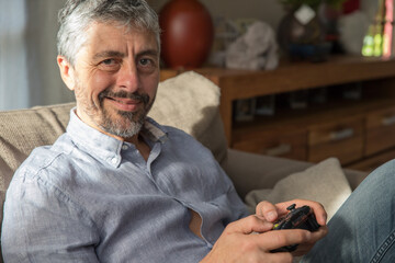 Un homme d'âge mur de 50 ans tient une manette de jeux vidéo dans ses mains et s'amuse en jouant à la console, il a le sourire - obrazy, fototapety, plakaty