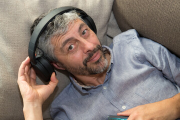 un homme d'âge mur 50 ans écoute de la musique trop fort avec son casque. Il l'enlève avec sa main et fait la grimace - obrazy, fototapety, plakaty