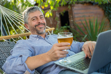 homme 50 ans allongé dans son hamac, boit une bière en travaillant avec son ordinateur