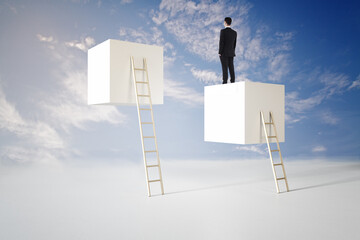 Back view of young businessman standing on abstract cubes with ladder on bright blue sky...
