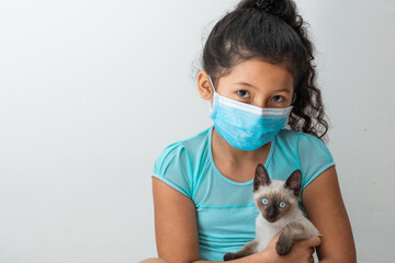 little girl (8 years old) sitting with a Siamese cat in her hands, brown girl with a blue surgical mask. Medical, pharmaceutical and sanitary concept.