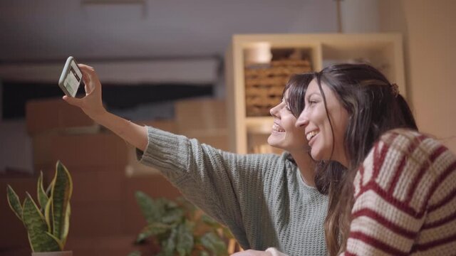 A female couple take a selfie in their new home during the relocation. Happy friends take pictures and use a smartphone.