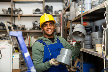 Cheerful Latino workman preparing for pipework routing, choosing supplies in shop of building...