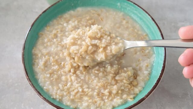 A hand stiring cooked oats porridge with a spoon in a green bowl, healthy breakfast and dieting product close up, tasty vegan food