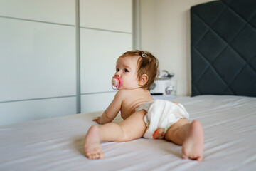 Obraz na płótnie Canvas Small caucasian baby girl six months old lying on the bed naked belly down with nipple pacifier wearing diaper at home in bright room copy space back view