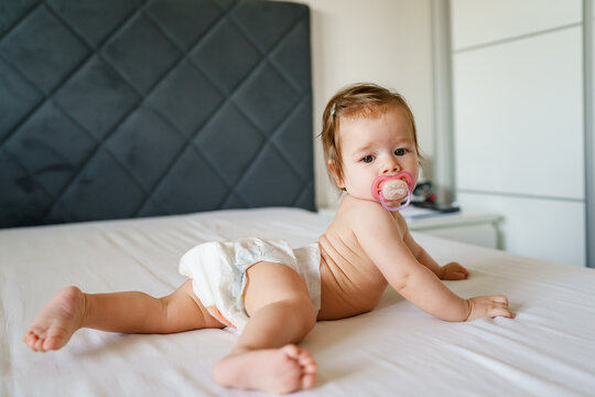 Small caucasian baby girl six months old lying on the bed naked belly down with nipple pacifier wearing diaper at home in bright room copy space