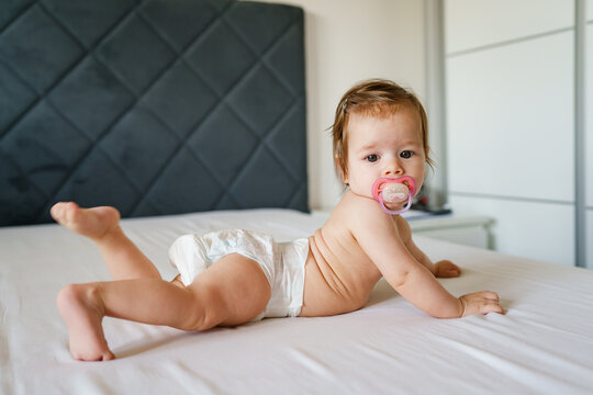 Small caucasian baby girl six months old lying on the bed naked belly down with nipple pacifier wearing diaper at home in bright room copy space side view