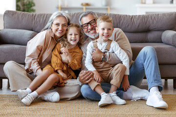 Happy family grandparents and two little kids embracing and smiling at camera while sitting on floor