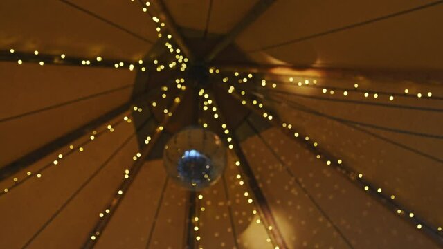 Party tent interior with disco ball hanging and reflecting light surrounded by LED fairy lights. 50 fps slow orbit and rack focus.