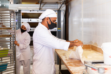 Bakery worker in protective mask cuts raw dough with a knife to prepare for baking