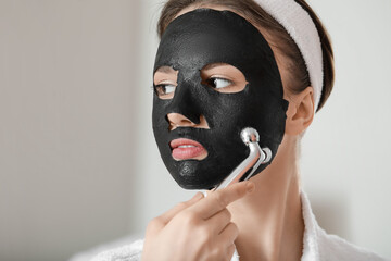 Beautiful young woman with sheet facial mask and massage tool at home