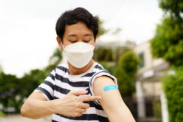 Young Asian man wearing covid-19 face protection mask pointing at bandage after receiving vaccine...