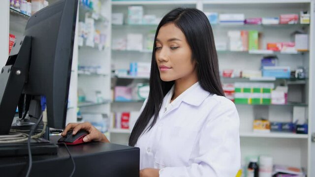 Doctor woman working in pharmacy store at hospital. Asian pharmacist female looking computer checking medication details in pharmacy drugstore. Concept of medical technology, business, lifestyle.