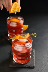 Woman squeezing fresh orange in glass with tasty Negroni cocktail on dark background