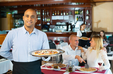 Fototapeta na wymiar Latino waiter with tray of delicious pizza on tray against the background of restaurant guests