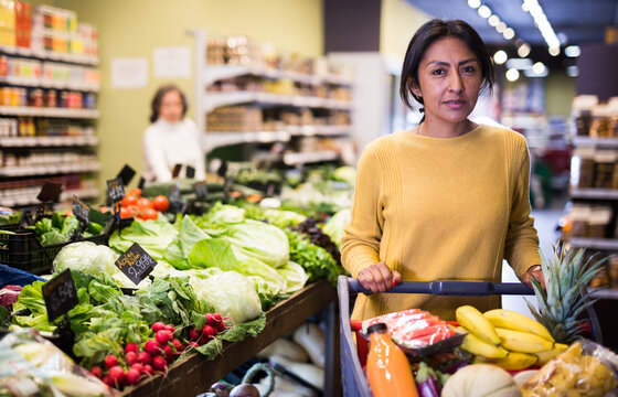 Portrait of satisfied hispanic woman with shopping trolley filled with fresh food goods bought in supermarket