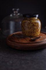 A silver spoon scooping from an open jar of whole grain mustard sitting on a wooden board on a black stone background. 