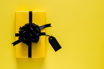 A yellow gift box with black ribbon and blank price tag puts on yellow background. Black friday and...