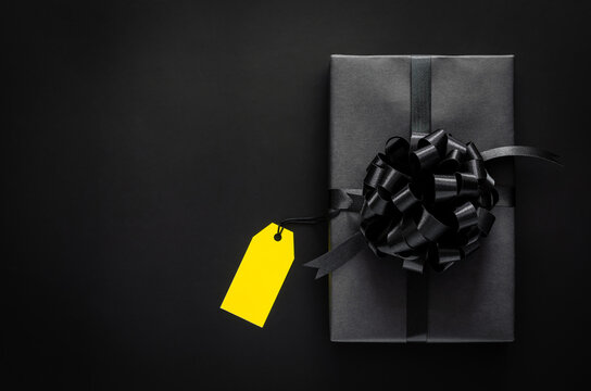 A Black Gift Box With Ribbon And Yellow Blank Price Tag Puts On Black Background. Black Friday And Boxing Day Concept.