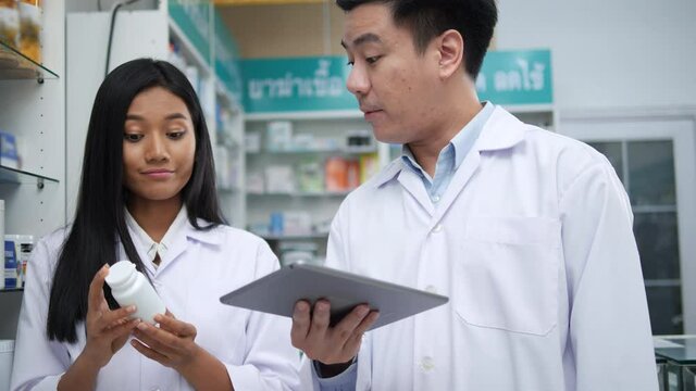 Asian pharmacists working and checking drug in pharmacy store at hospital. Asian doctor checking medicine cabinet and wearing white medical coat. Shelves pharmacy background in drugstore.