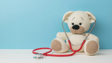 A beautiful knitted teddy bear with a stethoscope on a white table on a blue background.