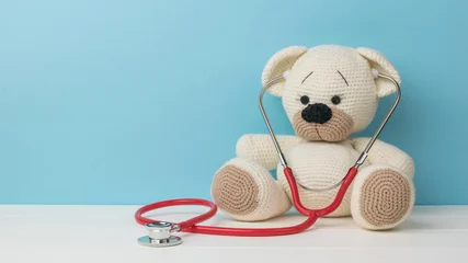 Fotobehang A beautiful knitted teddy bear with a stethoscope on a white table on a blue background. © kvladimirv