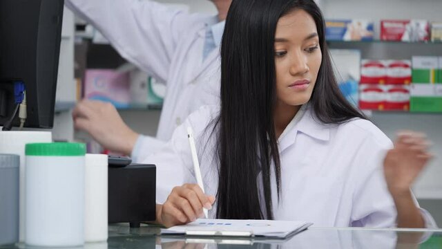 Asian pharmacists working and checking drug in pharmacy store at hospital. Asian doctor checking medicine cabinet and wearing white medical coat. Shelves pharmacy background in drugstore.