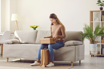 Happy young woman sitting on sofa at home unpacks cardboard parcel with online order from internet. Satisfied female customer opens box she got from package. Concept of delivery and online shopping.