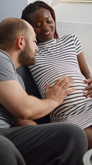 Fototapeta premium Married interracial couple bonding over child sitting at home. Young multi ethnic parents touching baby bump and talking about parenthood. Black pregnant woman and caucasian man
