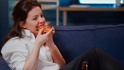 Young adult enjoying slice of pizza and beer sitting on sofa in living room while watching comedy on television. Woman eating fast food delivery, drinking beverage and laughing at TV