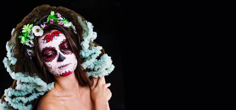 A young girl with dark hair and sugar skull makeup with flowers on her head looks into the camera on a black background. Panoramic stretched image for banner