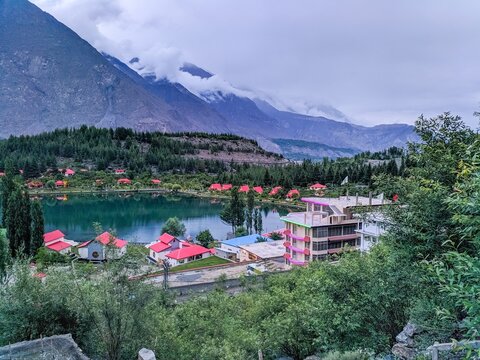 Stunning view of Kalam Valley (Swat, kpk) Pakistan showing restaurants with red roofs surrounding a scenic lake reflecting pines with pine trees and clouds hovering over high mountains in background