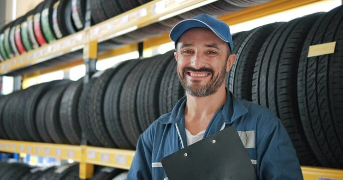 Portrait of smiling mechanic employee in work uniform looking camera at car tires shop service, Specialist tire fitting in the car service, checks the tire and rubber tread for safety.