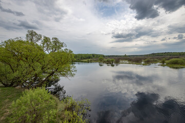 Naklejka premium Landscape with a dramatic sky reflected in the river. Early spring, juicy May greens. Bright green foliage on trees and bushes.