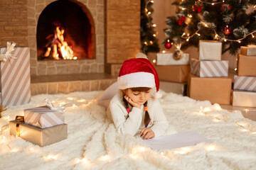 Charming little girl wearing white sweater and santa claus hat, lying on floor near Christmas tree, present boxes and fireplace, writing letter to Santa Claus.