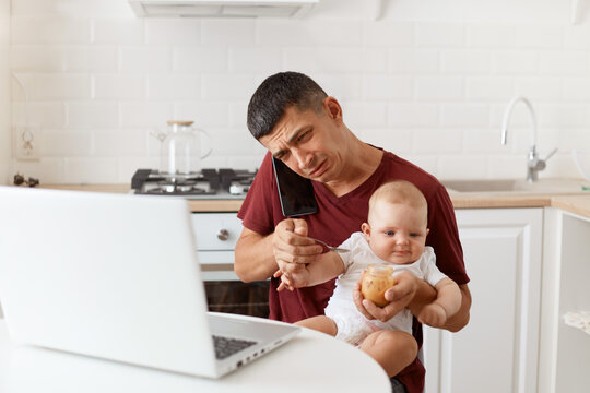 Stressed sad brunette male wearing maroon casual style t shirt sitting at table in kitchen, having conversation via smart phone, having sorrow look, feeding his infant daughter with fruit puree.