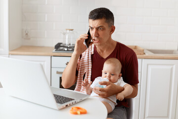 Scared serious man wearing burgundy casual t shirt with towel on his shoulder, looking after baby...