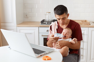 Indoor shot of man wearing burgundy casual t shirt with towel on his shoulder, looking after baby, giving crying daughter water from bottle, working online from home on laptop.