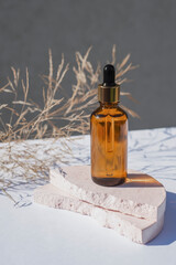 Amber glass dropper bottle with metallic lid on a podium with dry plants on light background. Skincare products , natural cosmetic. Beauty concept for face and body care