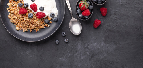 Horizontal banner with homemade granola, yogurt and fresh berries in black ceramic plate on dark stone background. Healthy vegan breakfast