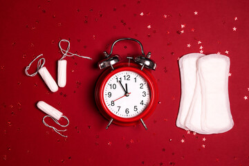 Menstrual pad and tampons with alarm clock and glitters on red background.