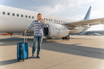 Happy passenger standing with suitcase and talking on mobile phone in airport. Trip concept