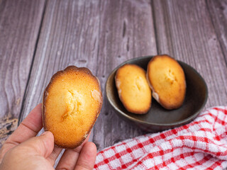 Hand holding a piece of madeleine on lemon glaze with madeleines on a dish and a cloth on a wooden table. Madeleine homemade traditional French small cookie. French confectionery concept
