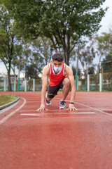 Young latin guy with protective mask, in position to run at full speed on a track, ready, at the start line, copy space