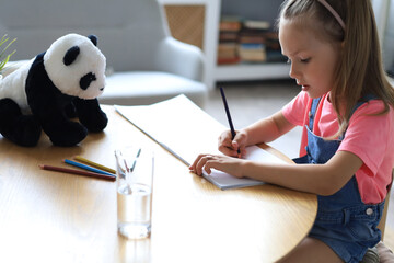 Smilling happy girl sitting in the table with a toy panda bear near to her enjoying creative...