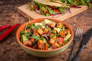Vegetarian salad with arugula, onions, tomatoes, avocado and fried tofu in a bowl on the table with tacos in the background, close-up. Modern vegetarian cuisine