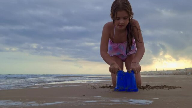 Low angle of beautiful caucasian little girl playing with bucket and sand on beach. Real-time